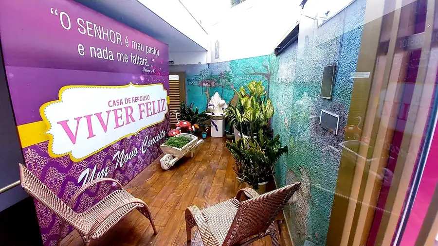 Jardim com piso de madeira, vasos de plantas, cadeiras de vime e painel com o logotipo Viver Feliz e a frase "O Senhor é meu pastor e nada me faltará"
