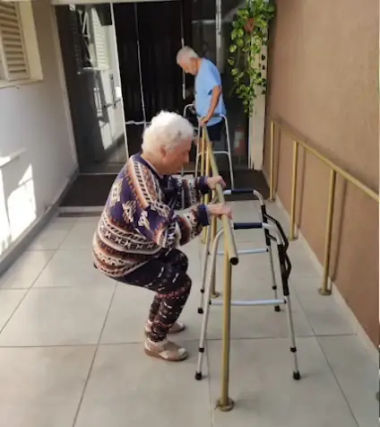 Moradora fazendo agachamento apoiada na barra do corredor, com andadores ao lado e outro morador ao fundo (quadro do vídeo)