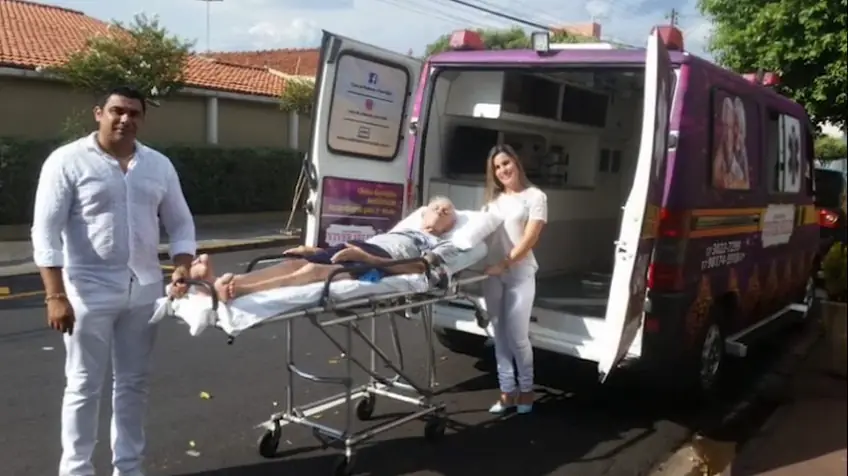 Um homem e uma mulher de branco ao lado da ambulância aberta, com uma pessoa deitada na maca (quadro do vídeo)