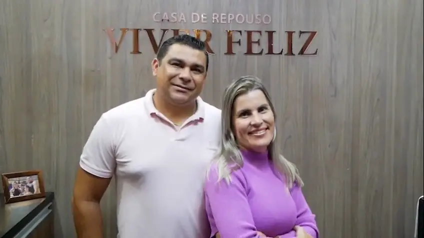 Um homem e uma mulher diante do painel de madeira com o letreiro Casa de Repouso Viver Feliz (quadro do vídeo)