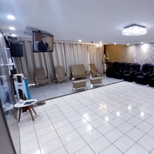 Salão com cadeiras de braço enfileiradas, TV na parede e lustre