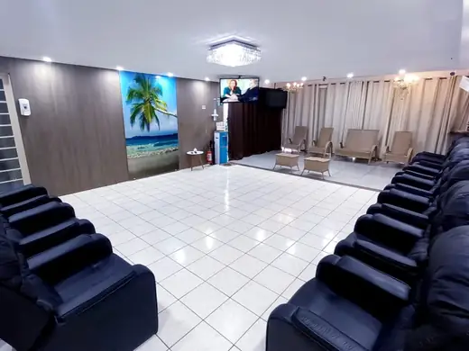 Salão com fileiras de poltronas reclináveis, painel de coqueiro na parede e cadeiras ao fundo