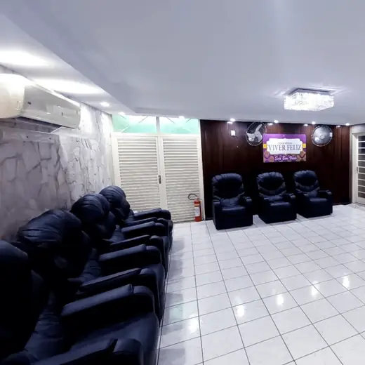 Salão de piso claro com poltronas reclináveis enfileiradas, lustre de cristal e TV