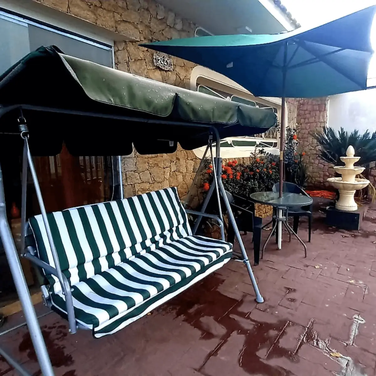 Varanda com balanço listrado de verde e branco sob toldo, mesa com guarda-sol e fonte de água